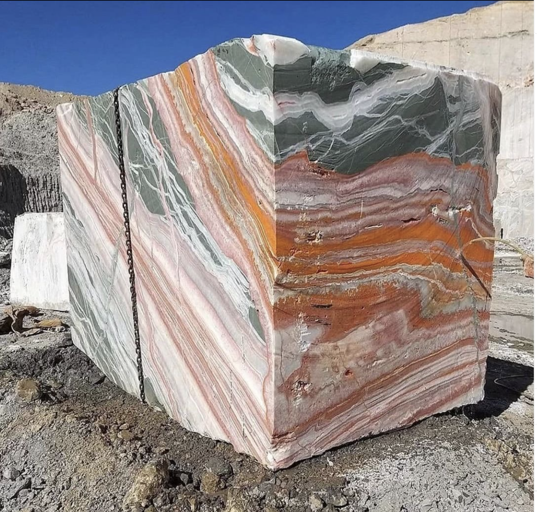 A massive raw block of Rainbow Onyx at a quarry, revealing the stunning natural layers of orange, green, and pink before being processed into slabs.