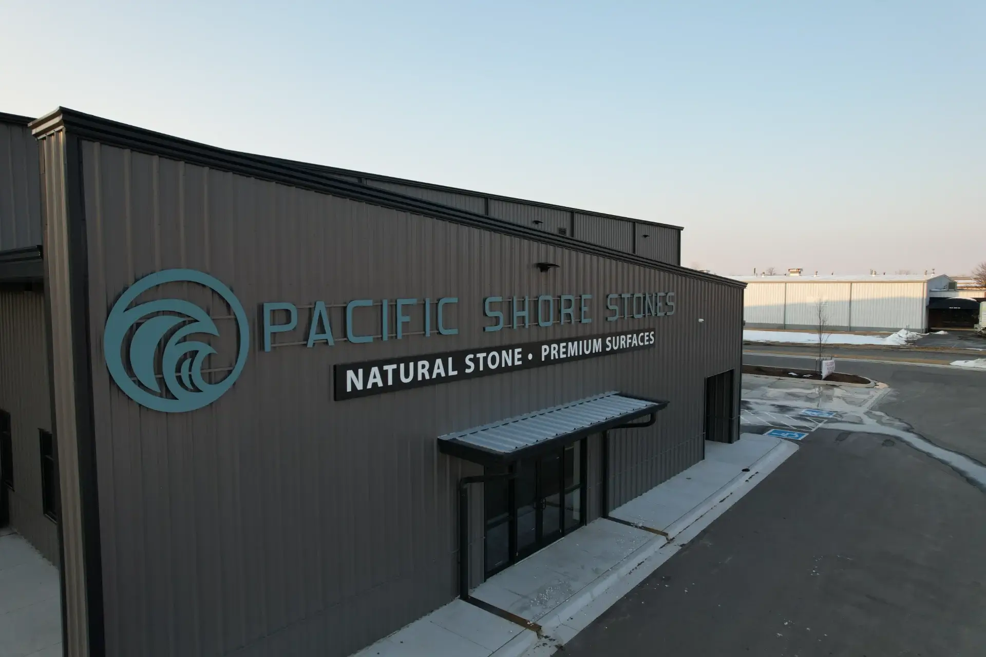 Exterior of Pacific Shore Stones building with teal logo and text 'Natural Stone Premium Surfaces' on grey siding