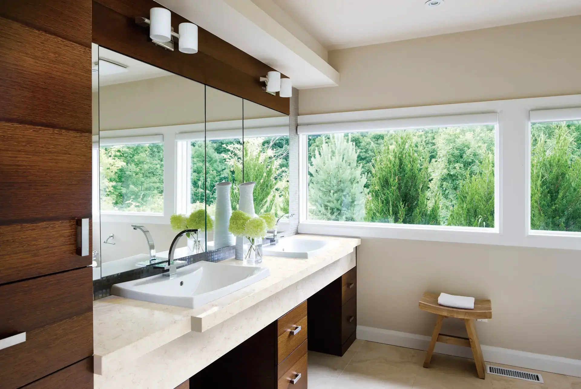 Modern bathroom with dual sinks, Clarino quartz countertop, wood vanity, large mirrored cabinets, and windows overlooking trees.