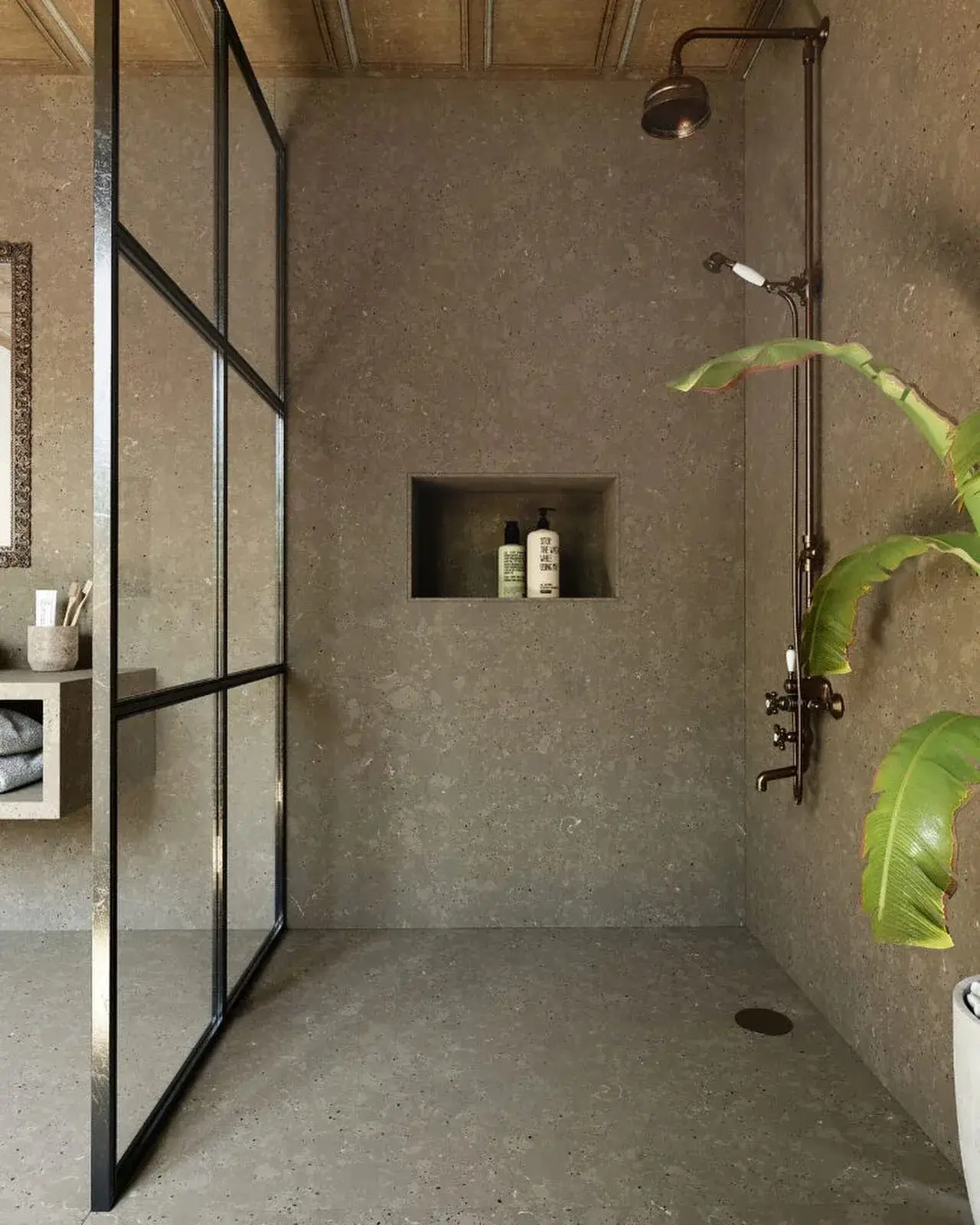Walk-in shower with dark gray terrazzo walls and floor, black-framed glass partition, bronze rainfall shower, and a large green plant.