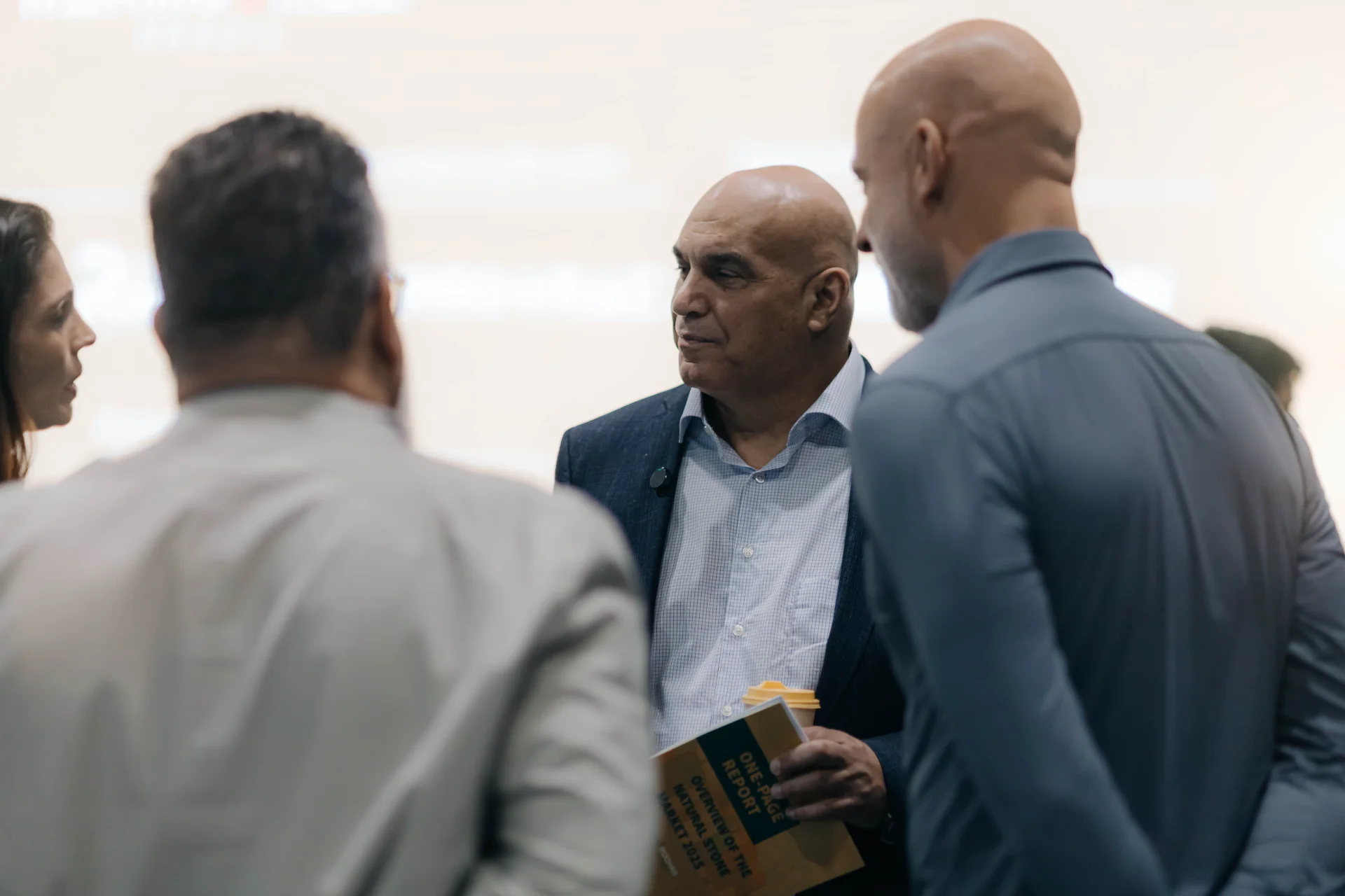 Bald man in blue suit jacket holding a yellow cup and report titled "ONE-PAGE REPORT OVERVIEW OF THE NATURAL STONE MARKET 2025" talking with two other people (one man in white shirt and one in blue shirt) at an indoor event, business casual