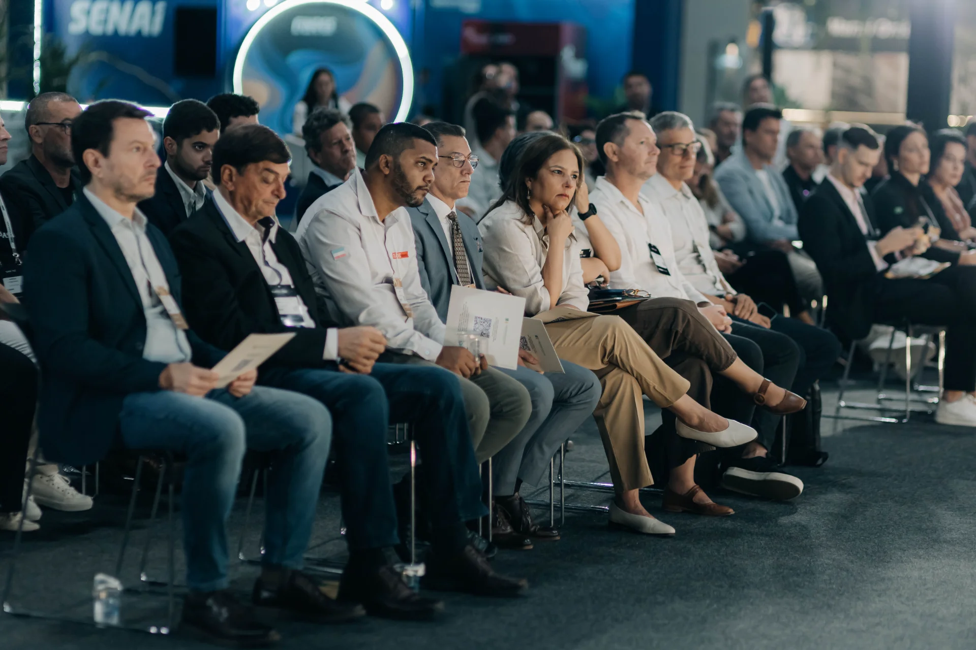 Diverse audience sitting attentively in rows at a conference or seminar, a woman in the foreground looks engaged.