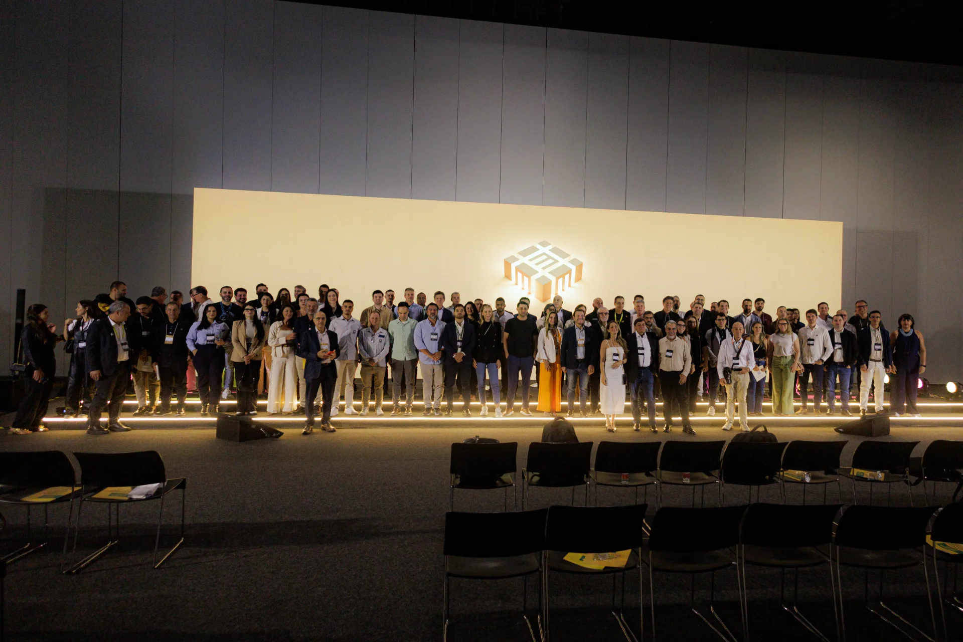 Large group of people standing on a brightly lit stage in front of a white screen with a 3D block logo.