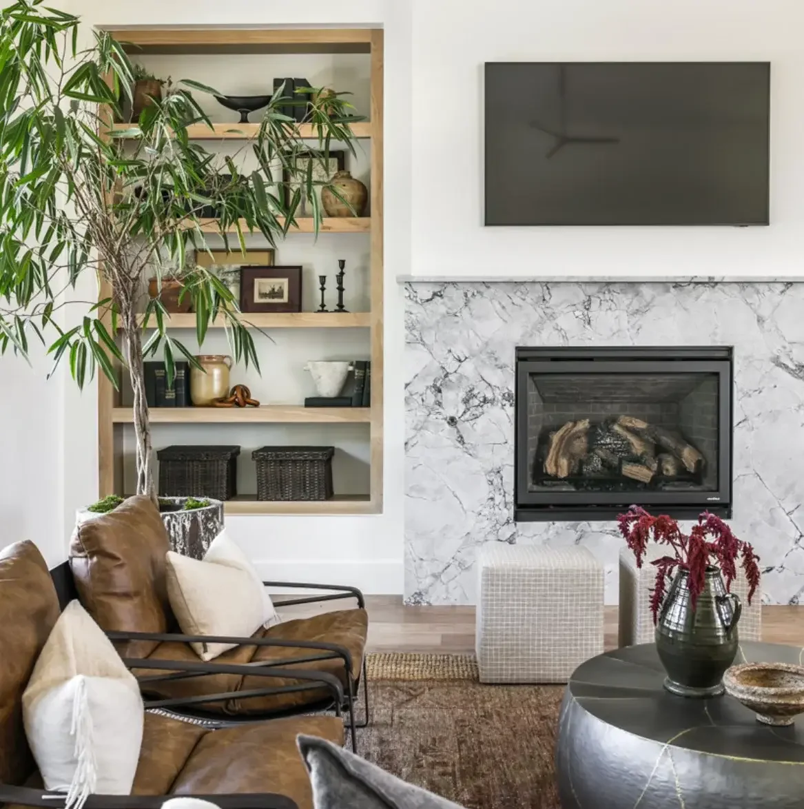 Modern living room with brown leather chairs, marble fireplace, black TV, built-in shelves, and a large plant.