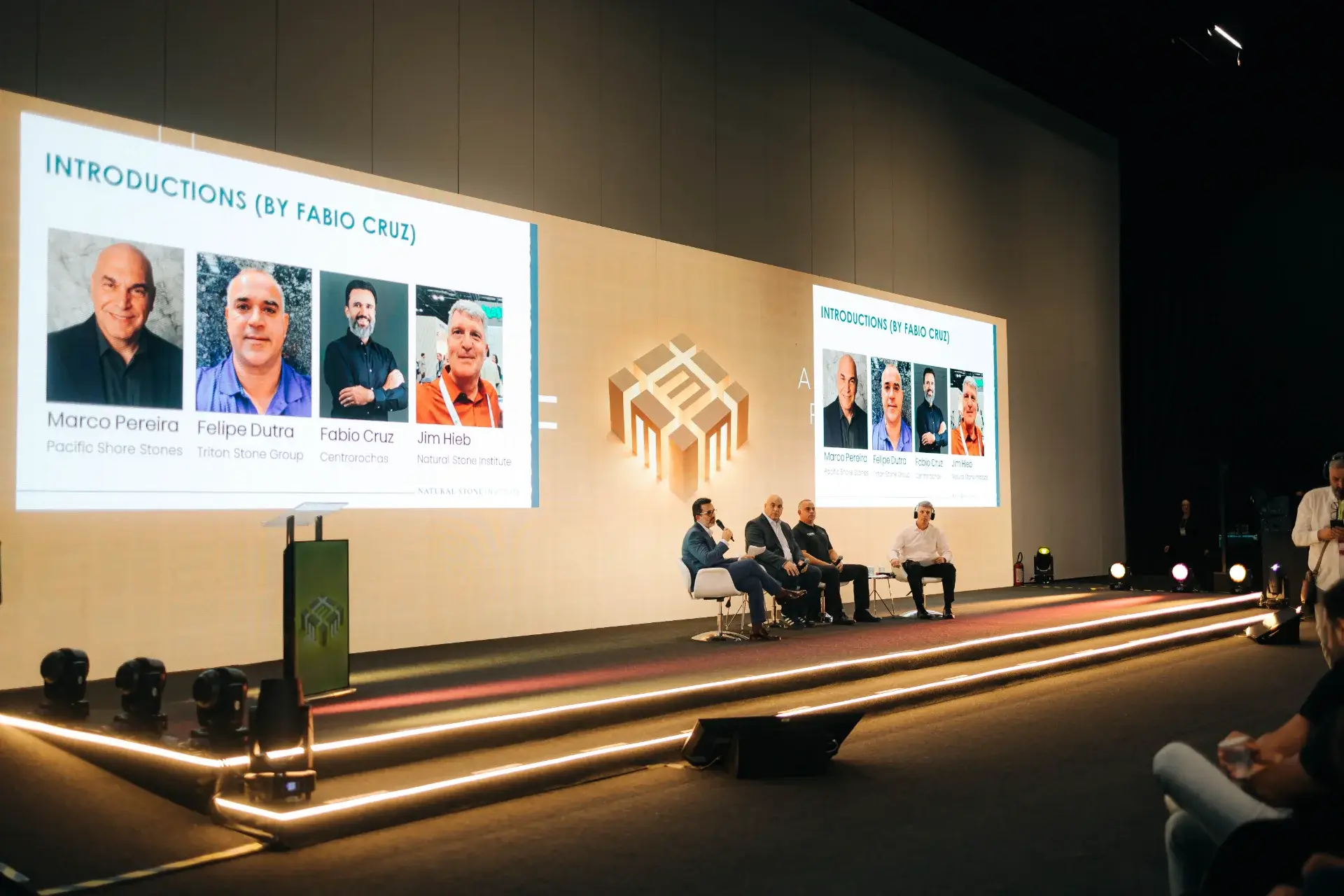 Four men in business attire sit on stage during a presentation by Fabio Cruz at the Natural Stone Institute event.