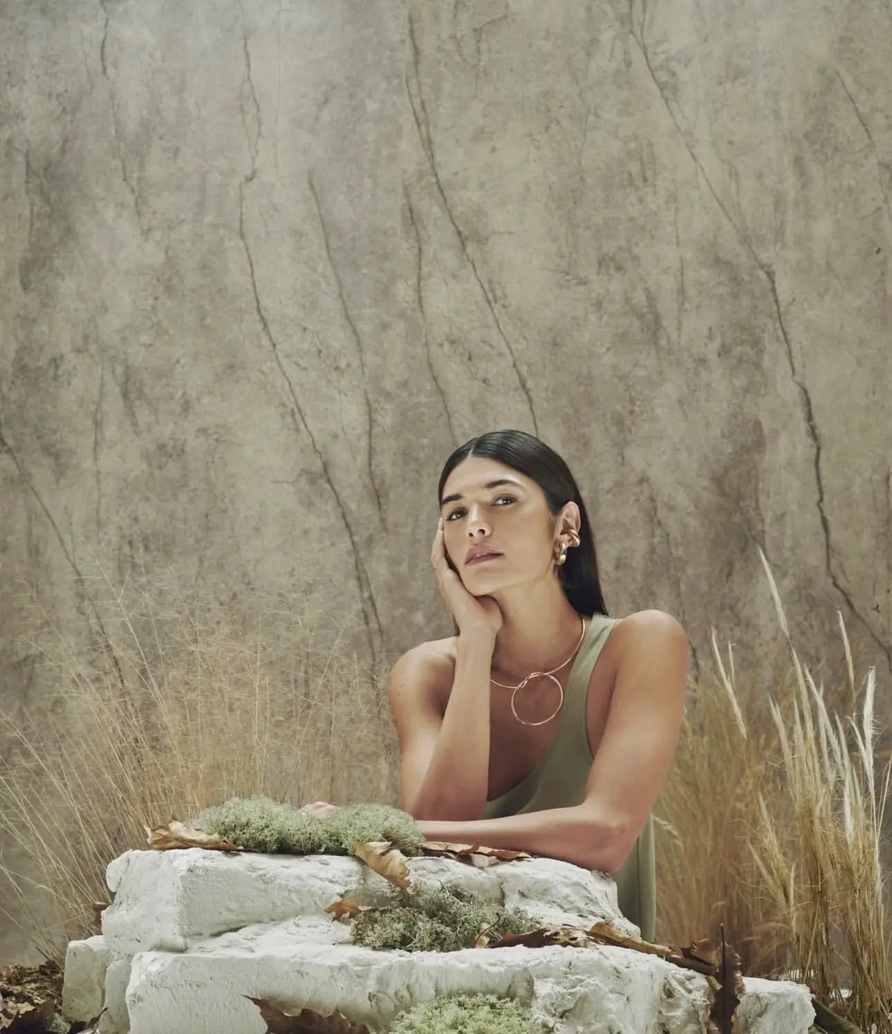 Woman wearing elegant gold jewelry: a circular necklace and statement earrings, against a natural stone wall and dried grass.