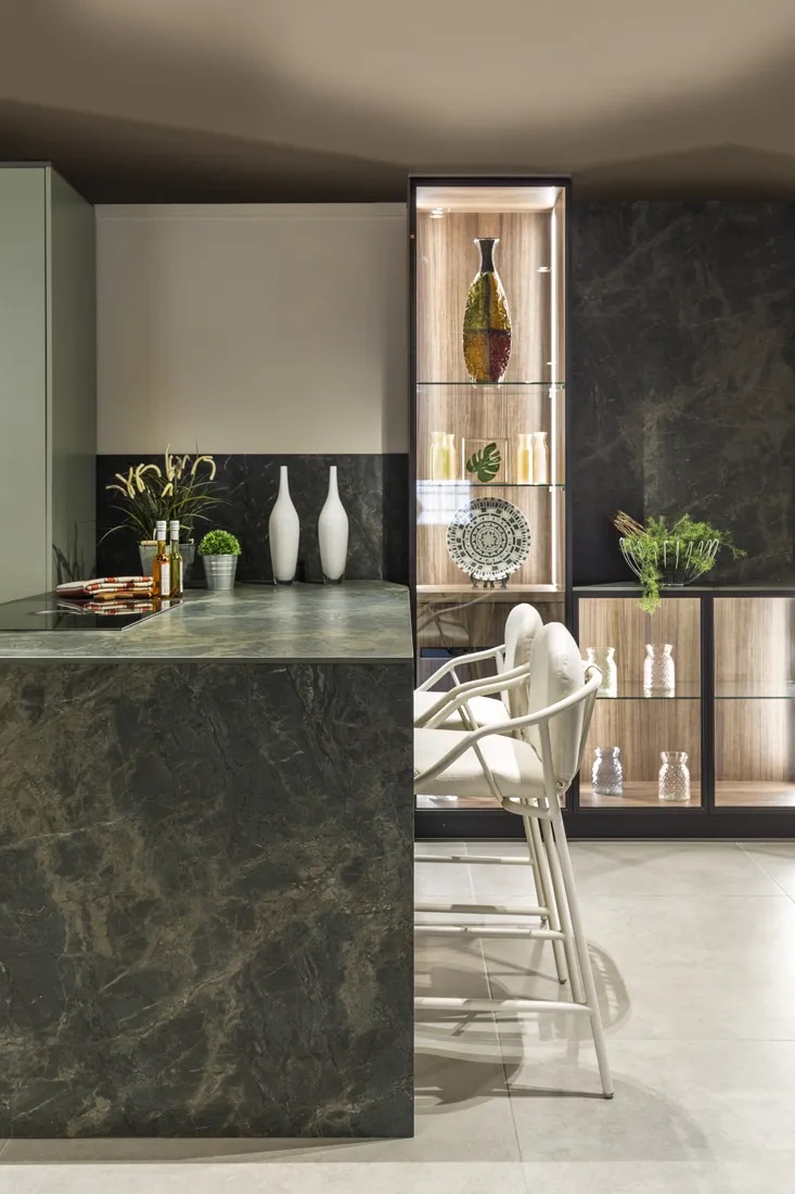 Modern kitchen island with dark marble countertop, two high-back stools, and display cabinets with decorative vases.