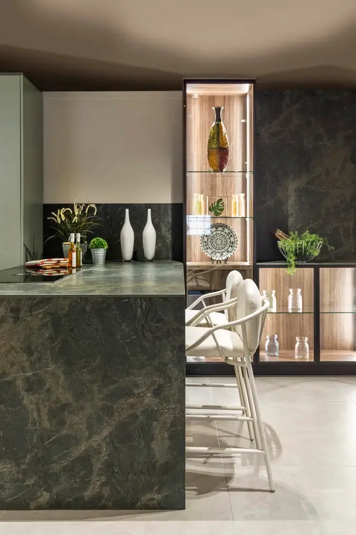 Modern kitchen island with dark marble countertop, two high-back stools, and display cabinets with decorative vases.