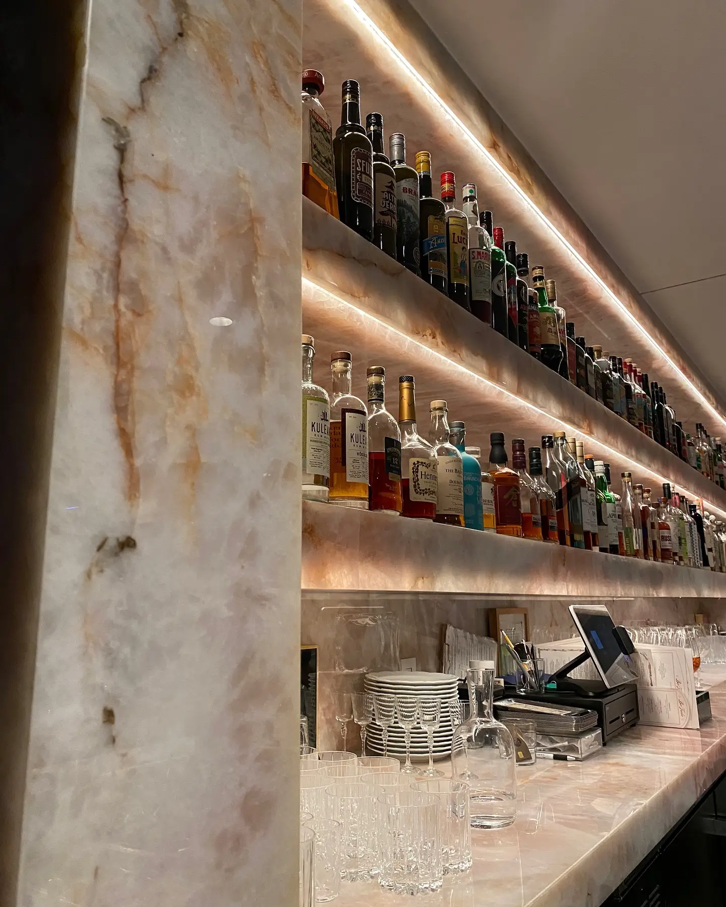 Well-stocked bar with glowing marble shelves displaying liquor bottles and shiny glassware.