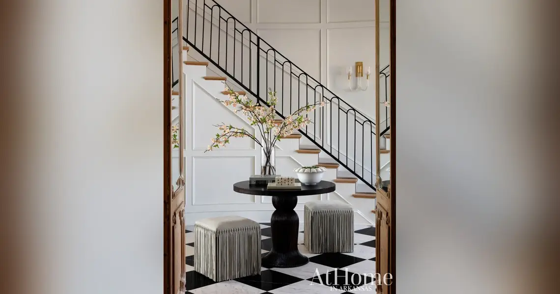 An elegant home entryway featuring black and white checkered marble flooring. In the center, a dark round pedestal table holds a clear vase filled with blooming pink and white cherry blossom branches.