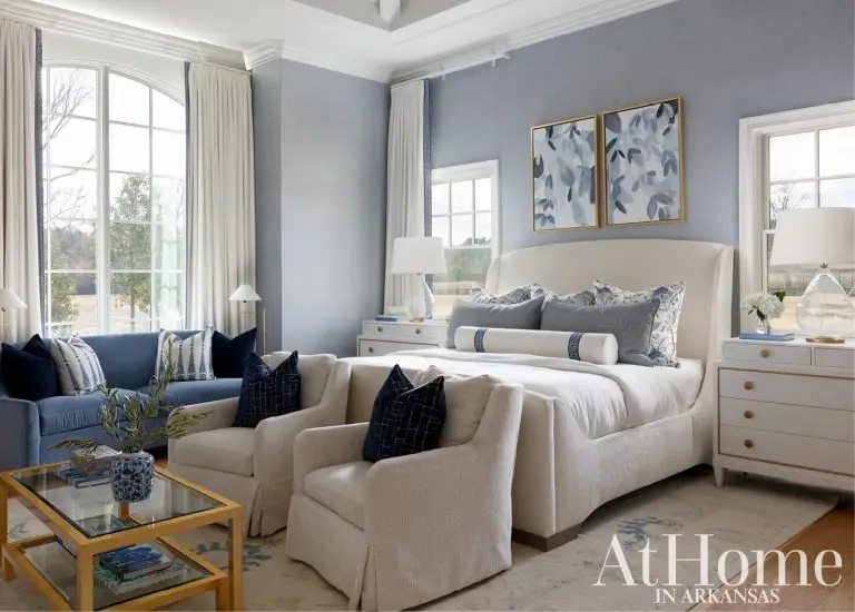 large master bedroom styled in shades of blue and cream. The bed has a curved cream upholstered headboard styled with navy and patterned accent pillows.