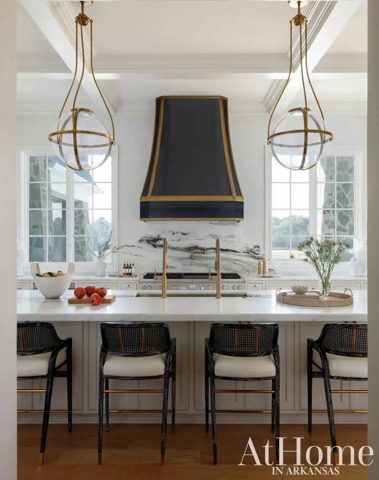 A kitchen island topped with white marble featuring dual brass gooseneck faucets. The island is paired with four black wooden counter stools with woven cane backs and light seats.