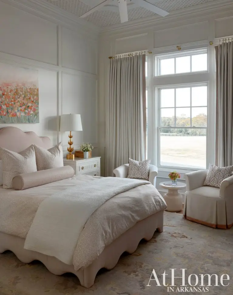 A bright and cozy bedroom decorated in neutral tones and soft blush pink. The bed features a scalloped upholstered headboard and base in pale pink, dressed in light linens and accent pillows.