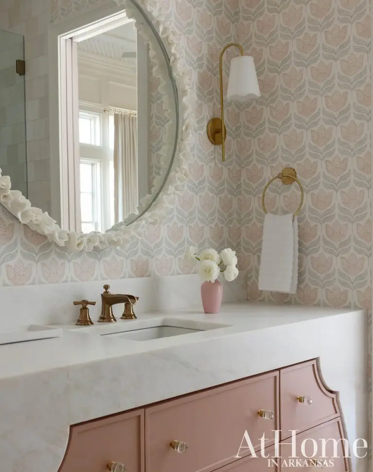 A soft and delicate bathroom wrapped in muted pink and gray floral pattern wallpaper. The white marble vanity features a vintage-style brass faucet set over a dusty-pink cabinet with glass knobs.