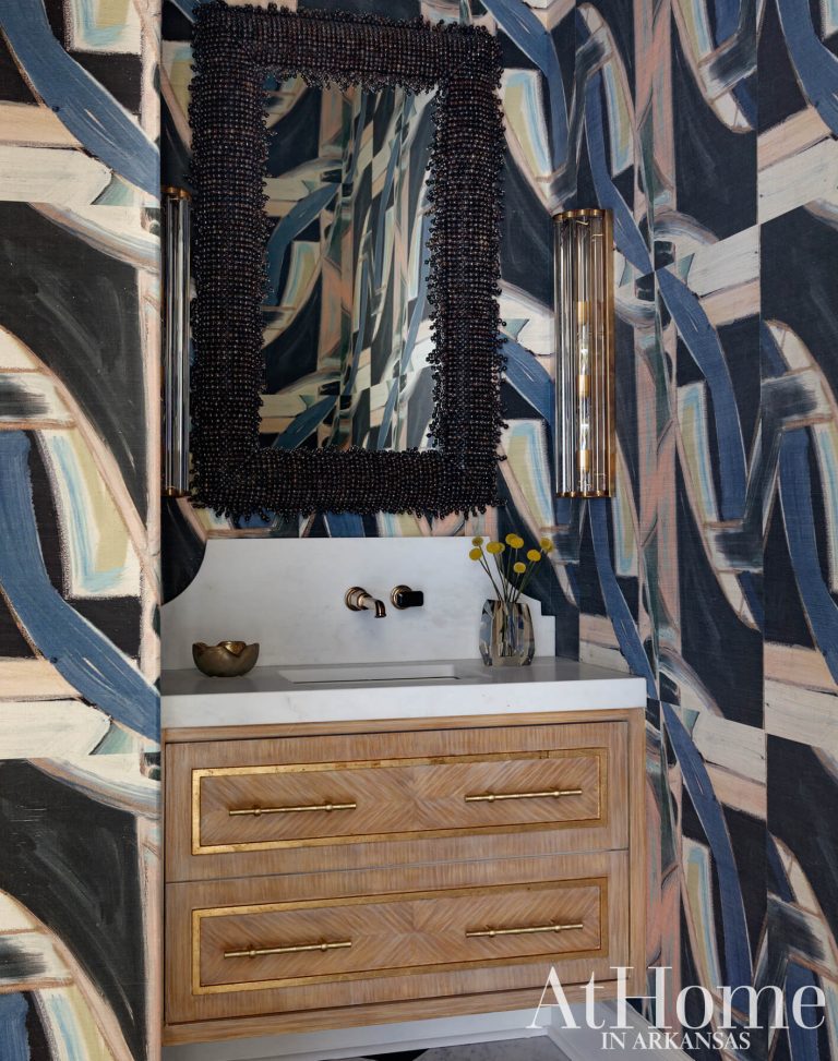 A modern powder room featuring abstract wallpaper with broad brushstrokes in shades of blue, beige, and black. A white marble countertop sits atop a light wood vanity accented with gold trim