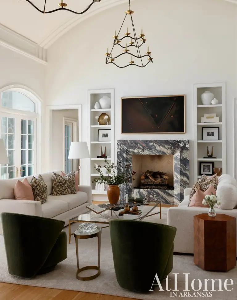 Elegant living room with high ceilings, two cream sofas facing a marble fireplace, white built-in bookshelves, and two dark green velvet swivel armchairs in the foreground.