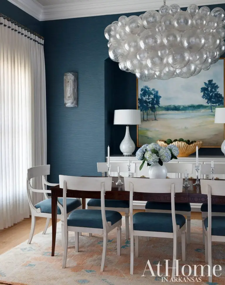 Elegant dining room featuring deep blue textured walls, a wooden table with white chairs, a large landscape painting, and a unique bubble glass chandelier.