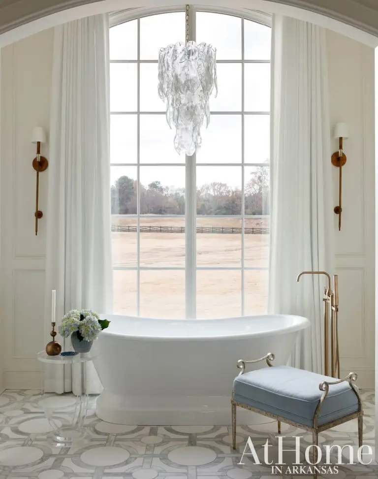 A spacious bathroom featuring a large arched window in the center that offers a view of an open field. In front of the window stands a white freestanding soaking tub.