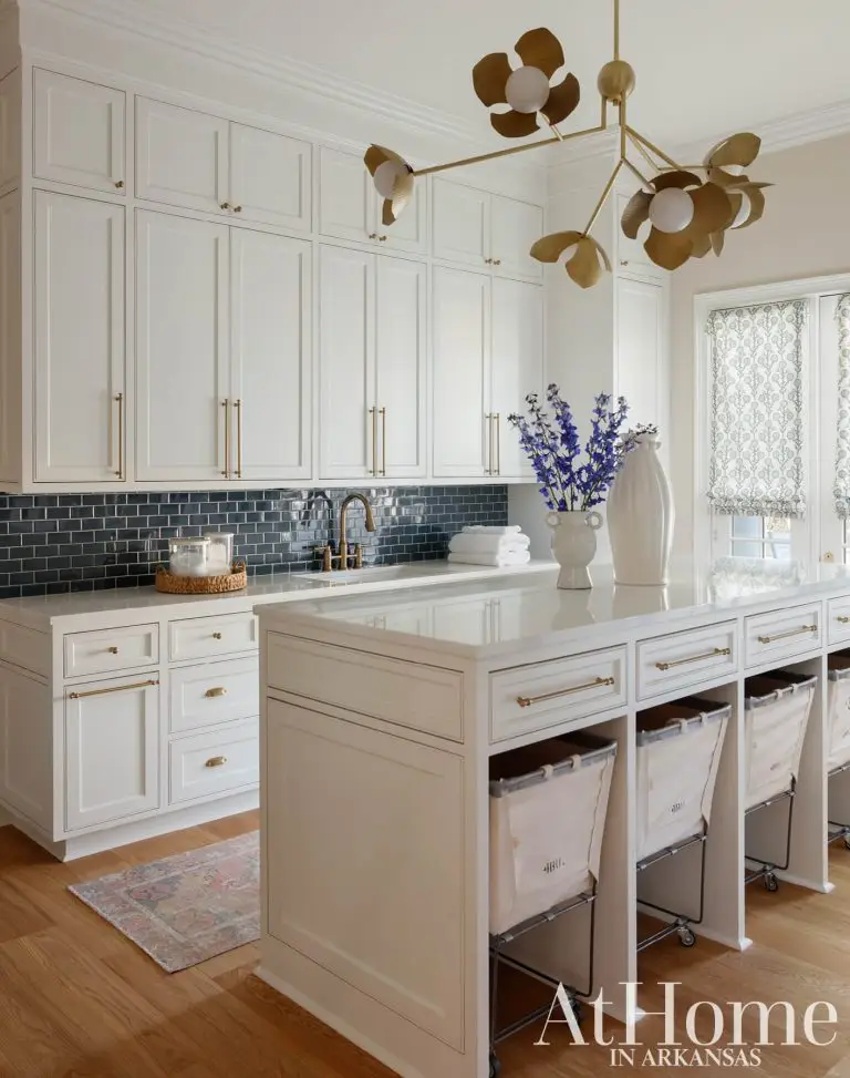 A spacious laundry room equipped with floor-to-ceiling white cabinets and brass hardware accents. The back counter features dark blue subway tile backsplash and a sink.