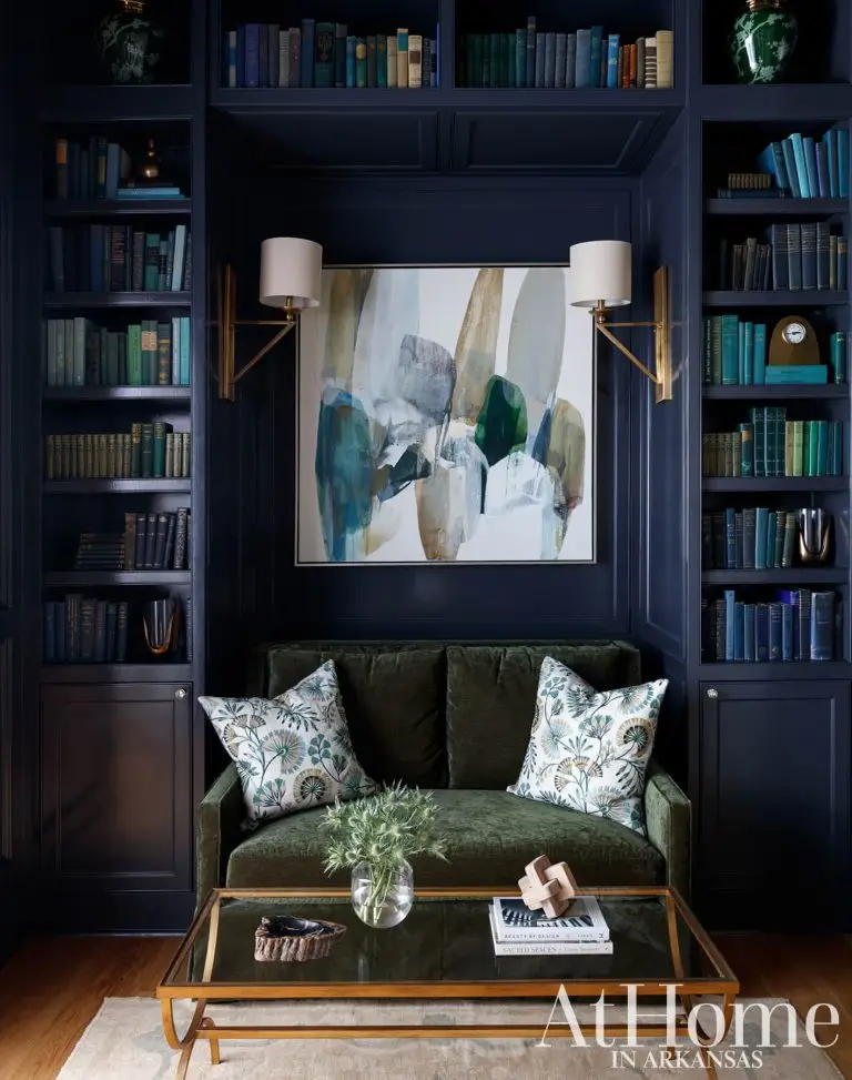 A moody and cozy reading nook framed by deep navy built-in bookcases filled with books. In the central recess sits a small olive-green velvet loveseat with patterned throw pillows.