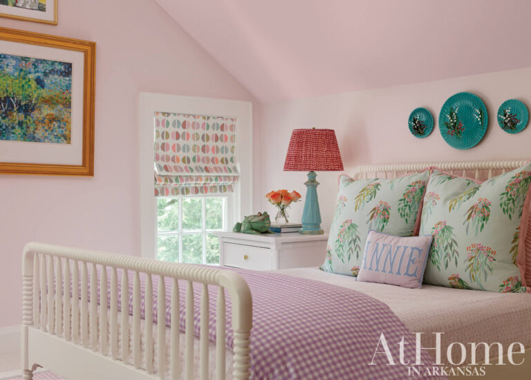 Pink bedroom with a white spindle bed, floral pillows, a red pleated lampshade, and teal Majolica plates hung above the bed.