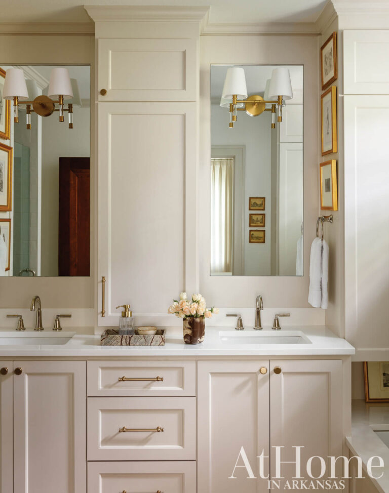 Primary bath with a white double vanity, framed mirrors, brass sconces, and framed prints on the walls.