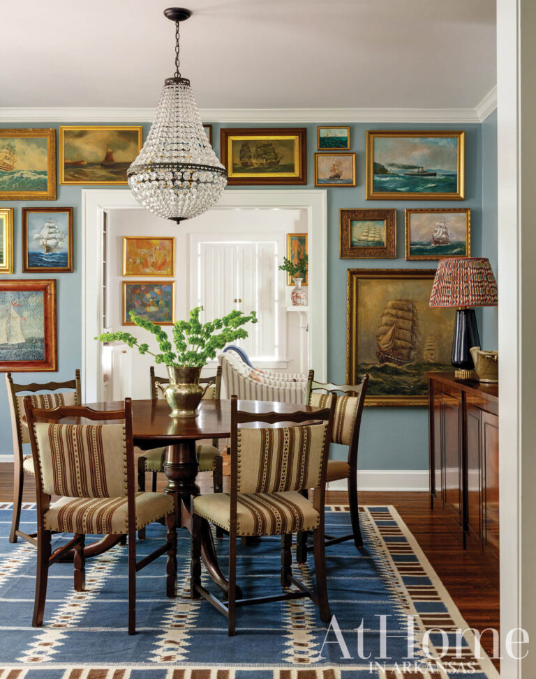 Dining room with blue walls hung with ship paintings, a crystal chandelier, a round wood table with striped chairs, and a blue geometric rug.