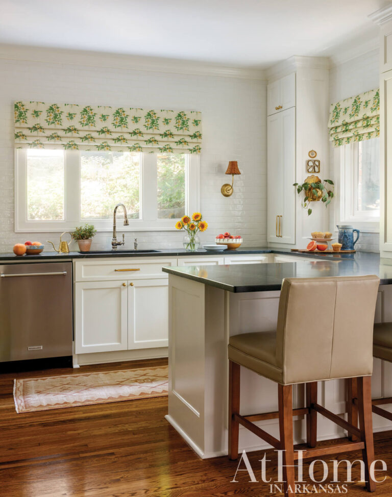 White kitchen with a dark countertop peninsula, beige leather barstools, green floral Roman shades, and stainless steel appliances.