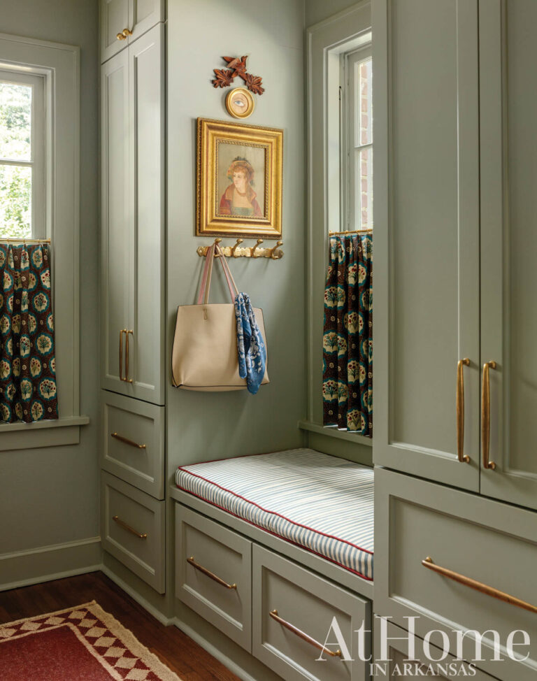 Green-gray pantry cabinetry with brass hardware, a built-in bench with a striped cushion, and a framed portrait above wall hooks.