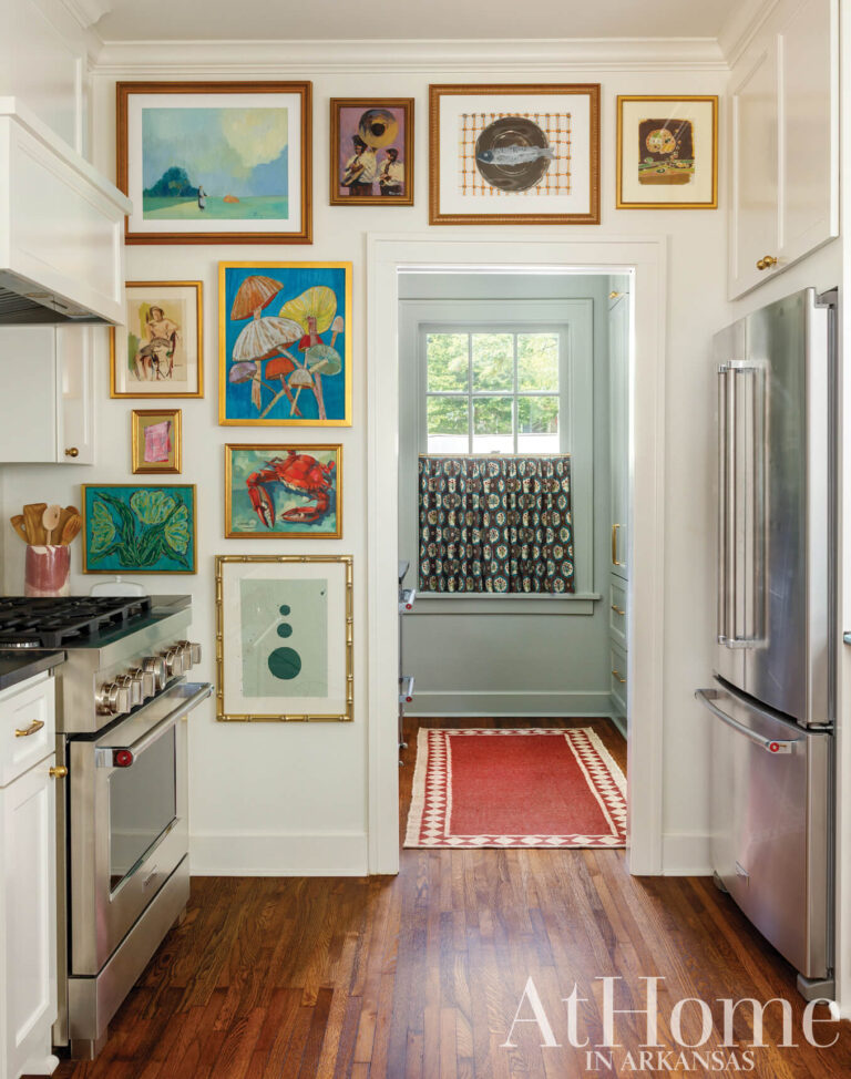 Kitchen wall of colorful framed art beside a stainless steel range and refrigerator, with a doorway leading into the pantry.