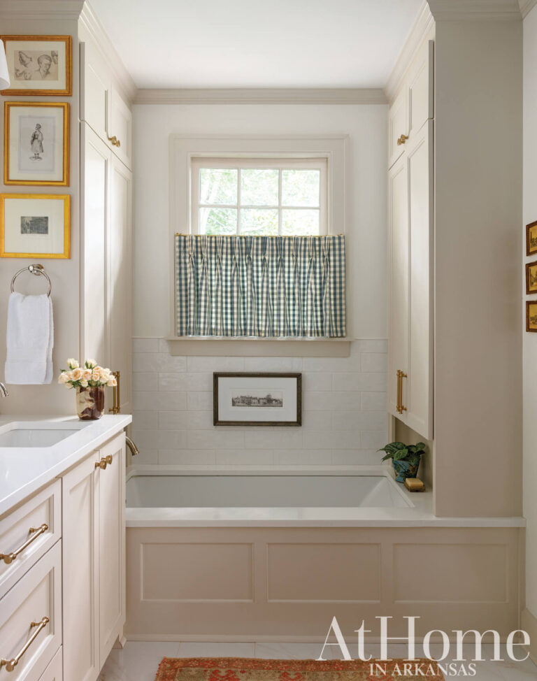 Primary bath with a paneled soaking tub beneath a window with a gingham café curtain, flanked by white built-in cabinets.