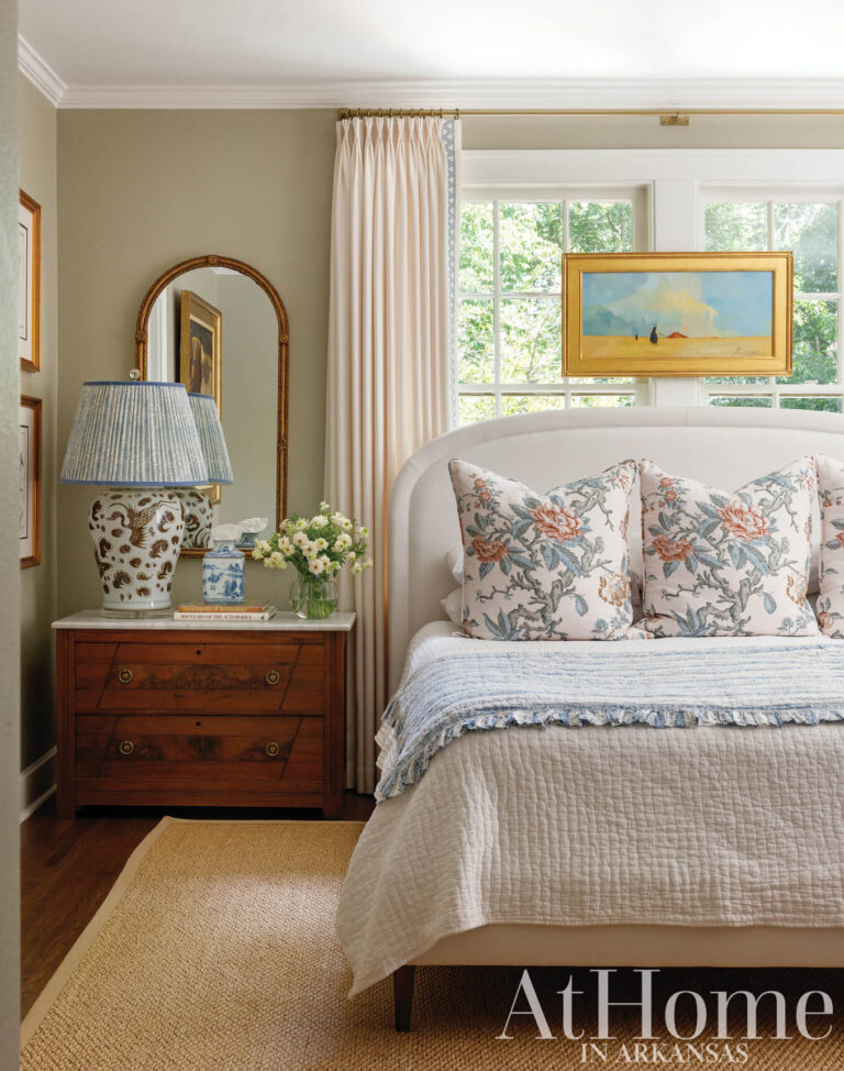 Primary bedroom with green-gray walls, an upholstered bed with floral pillows, a blue-and-white lamp on a wood chest, and a seascape painting.