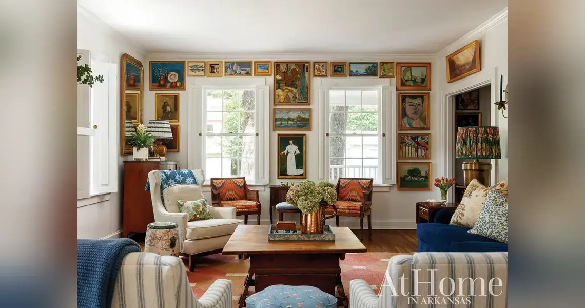 Living room with a gallery wall of framed paintings around the windows, flamestitch armchairs, a wood coffee table, and a patterned rug.