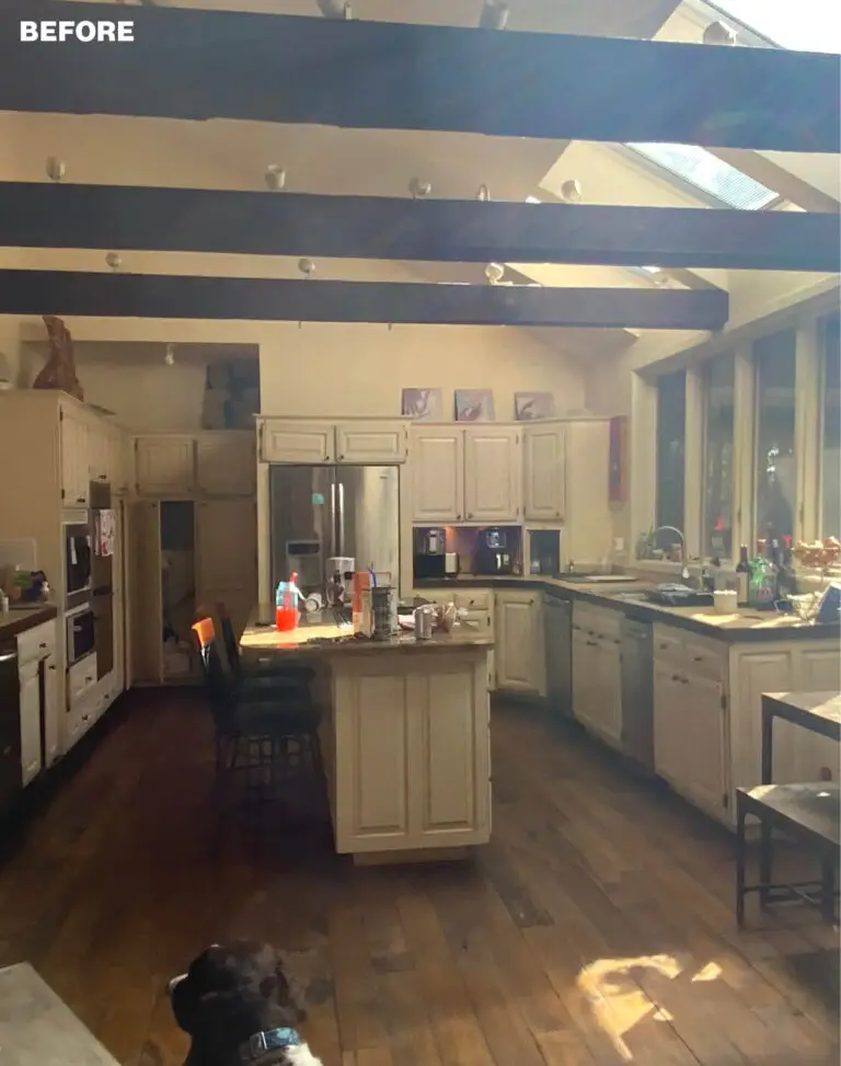 Before photo of the kitchen with cream distressed cabinets, a granite-topped island, dark ceiling beams, and wood floors.