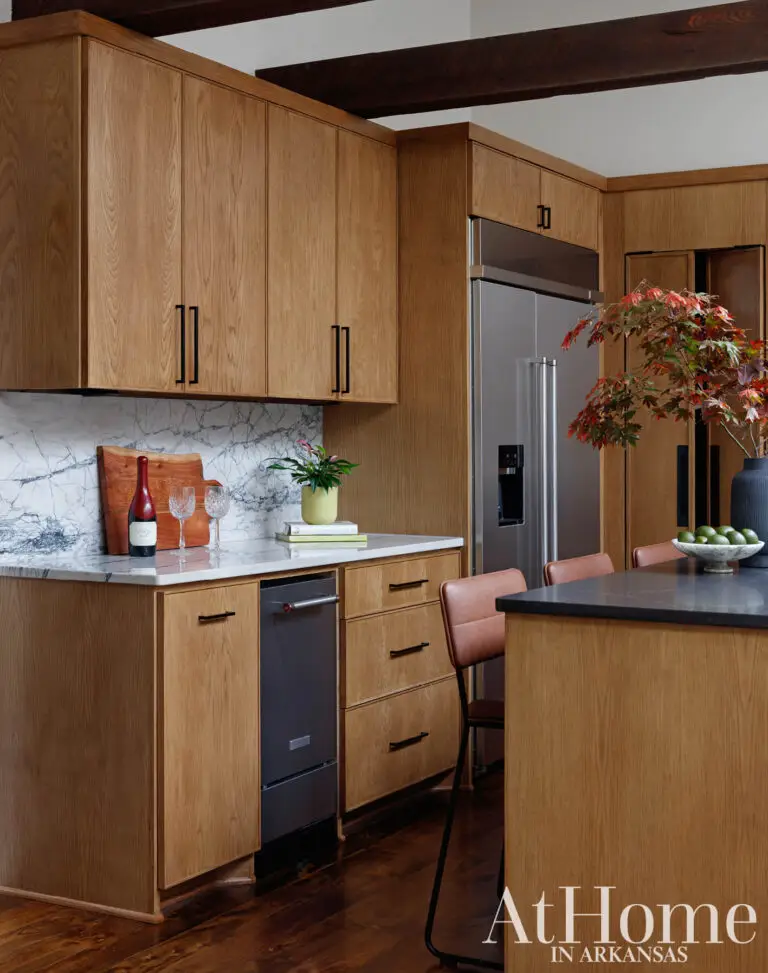 Stained oak cabinetry with a quartzite backsplash beverage station, a stainless steel refrigerator, and leather barstools at a dark-topped island.
